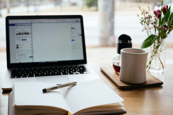 Foto av ei skrivebok og penn foran ein laptop, med eit te-krus ved sida av